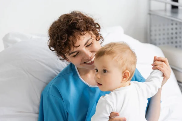 Pleased woman in patient gown holding hand of little daughter on bed in clinic — Stock Photo