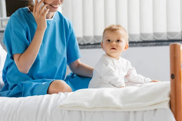 Toddler girl in romper sitting on hospital bed near smiling mom in patient gown talking on mobile phone — Stock Photo