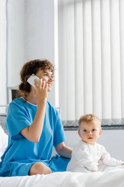 Happy woman in patient gown talking on smartphone near toddler child sitting on bed in clinic — Stock Photo