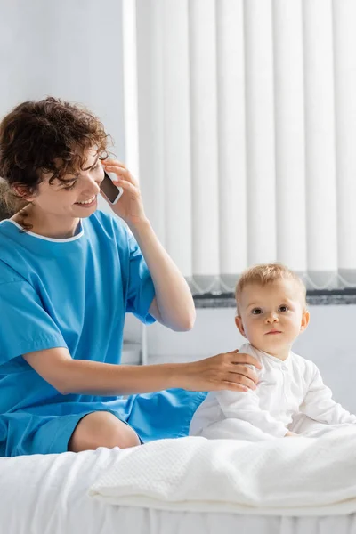 Smiling woman in patient gown touching baby while talking on cellphone on hospital bed — Stock Photo