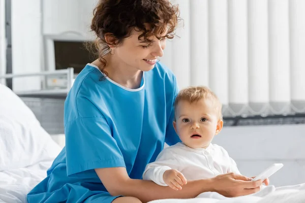 Happy woman in patient gown messaging on mobile phone near toddler daughter on bed in clinic — Stock Photo