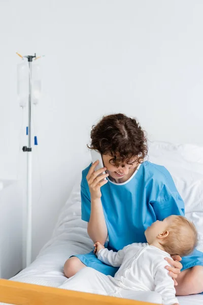Happy woman talking on cellphone while sitting on hospital bed near little daughter — Stock Photo