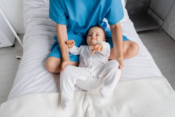 High angle view of woman in patient gown sitting with crossed legs near baby in romper on hospital bed — Stock Photo