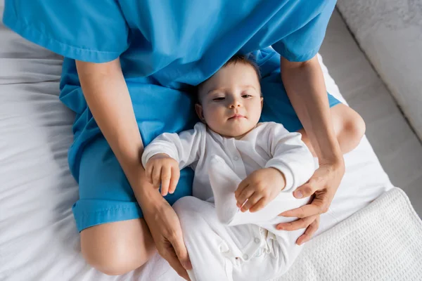 Top view of toddler child in romper near mother in patient gown sitting with crossed legs on hospital bed — Stock Photo