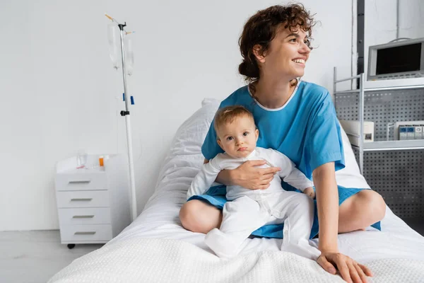 Cheerful woman in patient gown looking away while sitting with toddler baby on hospital bed — Stock Photo
