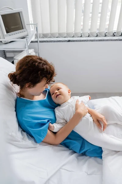 Toddler girl in romper smiling near mom in patient gown on bed in clinic — Stock Photo