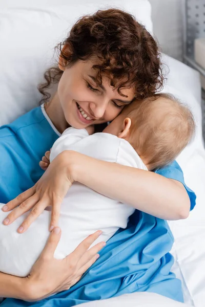 Happy woman with closed eyes embracing little child in romper on bed in hospital — Stock Photo