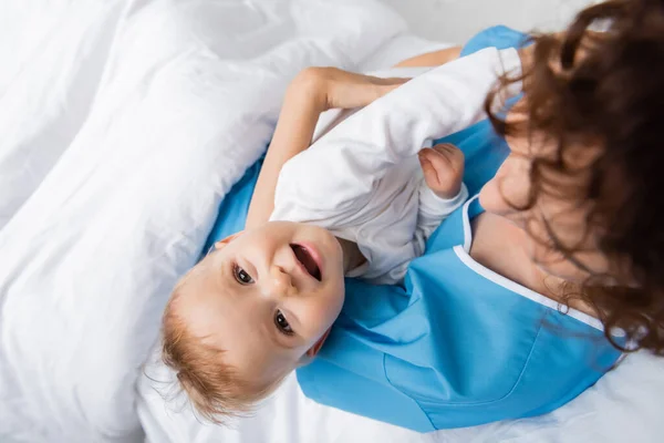 Top view of cheerful baby in romper smiling at camera near blurred mother on hospital bed — Stock Photo