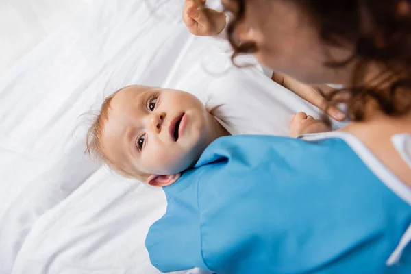 Top view of little child looking away while lying on hospital bed near blurred mom - foto de stock