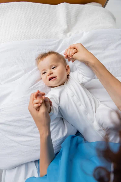 Top view of woman holding hands of little baby in romper smiling on bed in clinic — Stock Photo