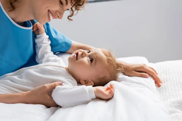 Side view of happy woman looking at little girl in romper lying on bed in hospital — Stock Photo