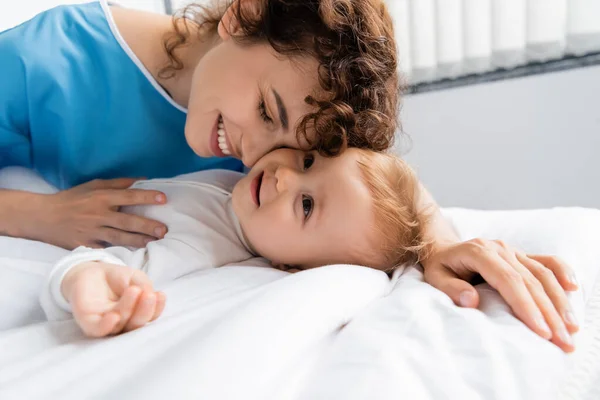 Smiling woman in patient gown embracing toddler child lying on bed in hospital ward — Stock Photo