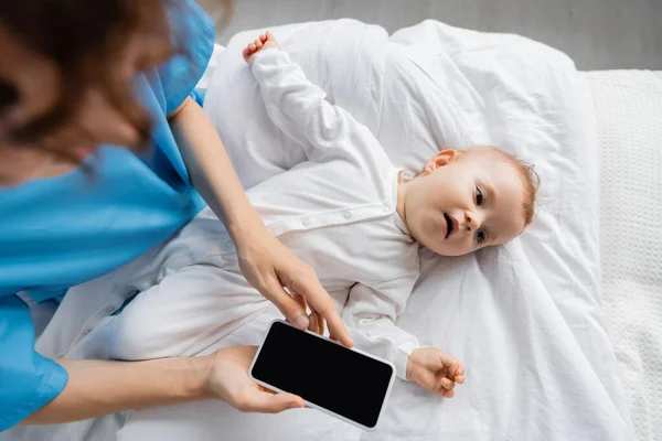 Top view of little child in romper lying on hospital bed near blurred mom holding smartphone with blank screen — Stock Photo