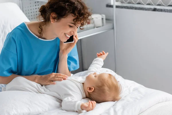 Pleased woman in patient gown looking at toddler daughter while talking on mobile phone in hospital ward — Photo de stock