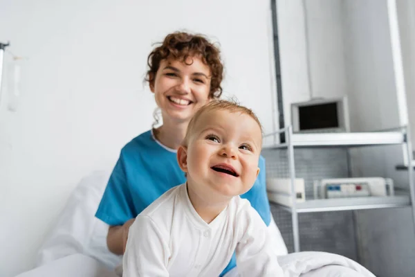 Cheerful baby in romper looking away near mom smiling on blurred background in hospital ward — Fotografia de Stock
