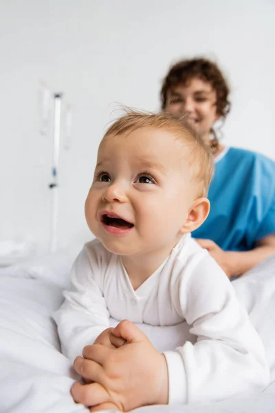 Cheerful baby in romper crawling on hospital bed near mother smiling on blurred background — Stock Photo