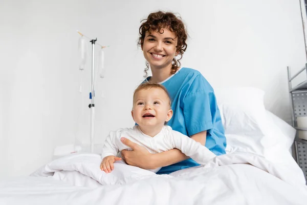 Cheerful woman looking at camera while sitting with smiling girl on bed in hospital ward — Stock Photo