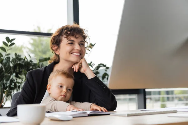 Smiling businesswoman looking at computer monitor while working in office near toddler daughter — Stock Photo