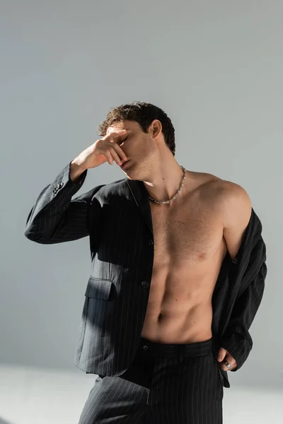 Shirtless man in black blazer and pants covering face with hand while standing on grey background - foto de stock