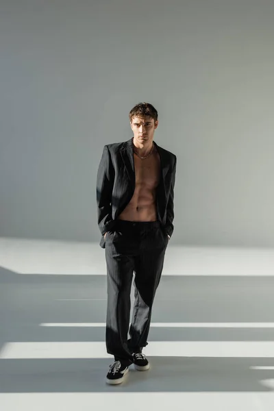 Full length of shirtless man in black suit and sneakers standing with hands in pockets on grey background with lighting — Stock Photo
