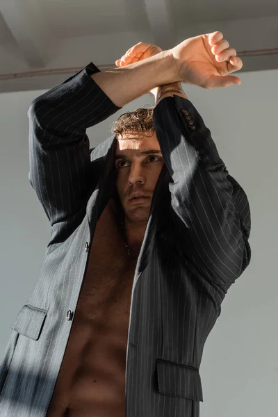 Shirtless man in black and striped blazer posing with crossed arms above head on grey background — Foto stock