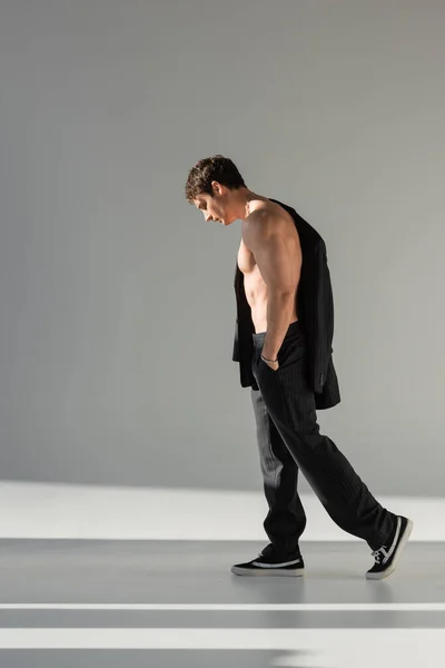 Full length of shirtless man with hand in pocket of black trousers walking in light on grey background — Stock Photo