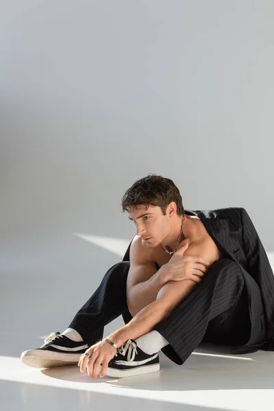 Sexy man with muscular body sitting in pants and sneakers under black blazer while looking away on grey background — Fotografia de Stock