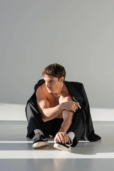 Shirtless muscular man in sneakers and pants sitting under black blazer and looking away on grey background — Fotografia de Stock