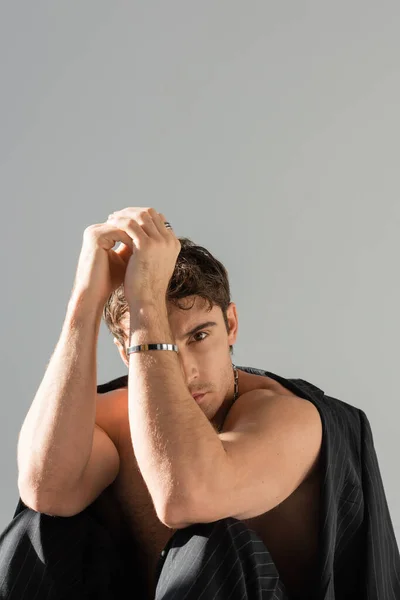 Shirtless man with muscular arms looking at camera while sitting under black blazer isolated on grey — Stock Photo