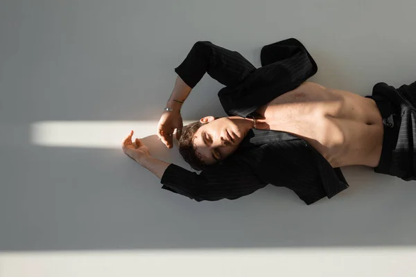 Вид зверху без сорочки чоловік у чорному блістері, що лежить із закритими очима на сонячному світлі на сірому фоні — Stock Photo