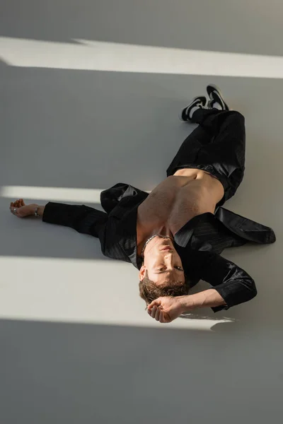 Высокий угол зрения человека в черных брюках и блейзере на тело без рубашки, лежащее в солнечном свете на сером фоне — стоковое фото