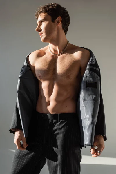 Sexy and muscular man looking away while posing in pants and black blazer on shirtless body on grey background — Stock Photo