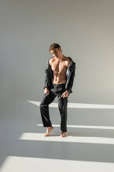 Полная длина босоногого человека с мускулистым туловищем, позирующим в черном пиджаке и брюках на сером фоне — стоковое фото