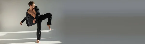 Full length of trendy barefoot man in black pants and blazer on shirtless body posing on one leg on grey background, banner — Fotografia de Stock