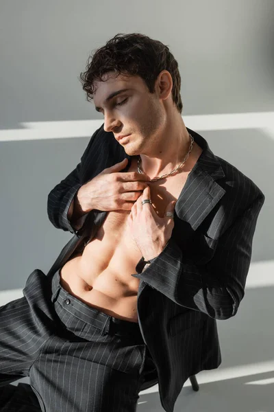 Man in silver necklace and black stylish blazer on shirtless torso sitting with closed eyes and hands near chest on grey background - foto de stock