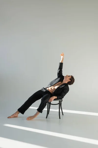 Full length of barefoot man in black suit posing on chair with raised hand on grey background with copy space — Stockfoto