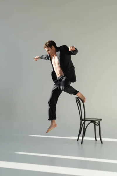 Повна довжина м'язистого чоловіка в чорному костюмі, що стрибає зі стільця з витягнутими руками на сірому фоні — стокове фото