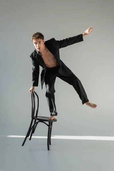 Full length of trendy barefoot man in black suit looking at camera while balancing on chair on grey background - foto de stock