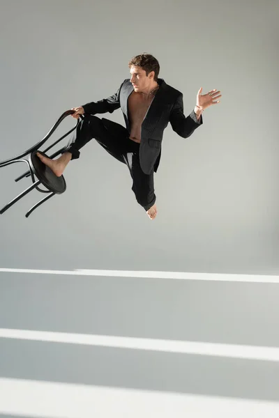 Повнометражний босоніж чоловік у модному костюмі, стрибає зі стільцем на сірому фоні з освітленням — стокове фото
