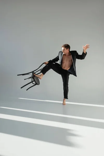Повнометражний босоніж і без сорочки чоловік в чорному стильному костюмі позує зі стільцем на сірому фоні — стокове фото