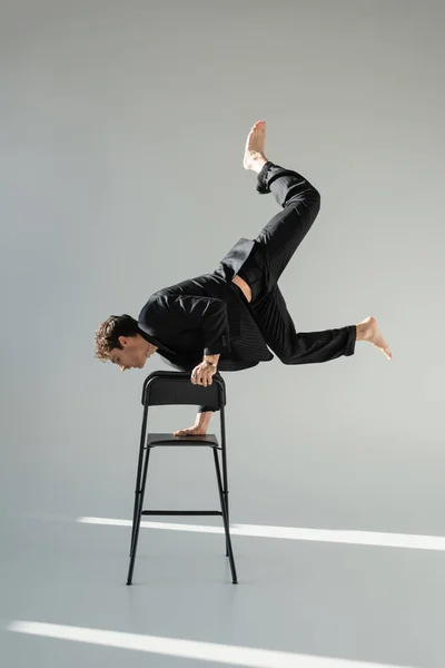 Full length of trendy barefoot man doing handstand on chair on grey background — Stockfoto