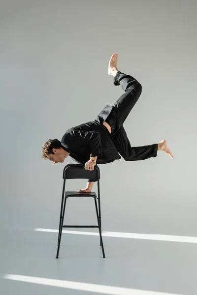 Full length of trendy barefoot man doing handstand on chair on grey background — Fotografia de Stock