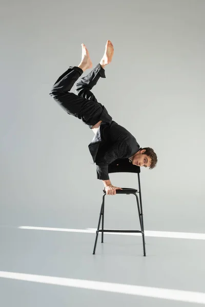 Full length of barefoot man in black suit doing handstand on chair and looking at camera on grey background — Stock Photo