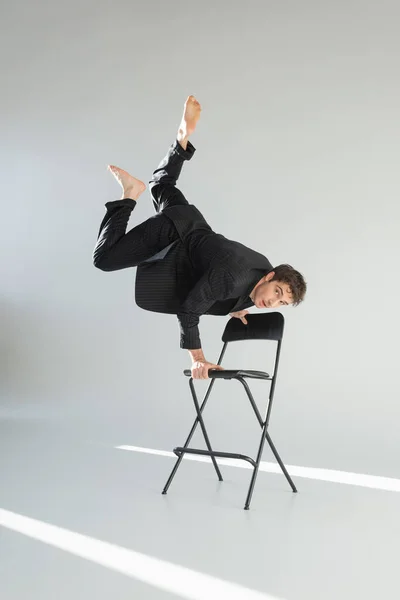 Full length of barefoot man in black trendy suit looking at camera and doing handstand on chair on grey background — Fotografia de Stock