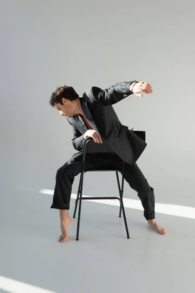 Повнометражний босоніж чоловік в модному блістері позує на стільці на сірому фоні з освітленням — стокове фото