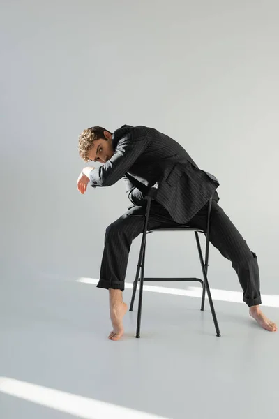 Full length of barefoot man in black and striped suit posing on chair and looking at camera on grey background — Stockfoto