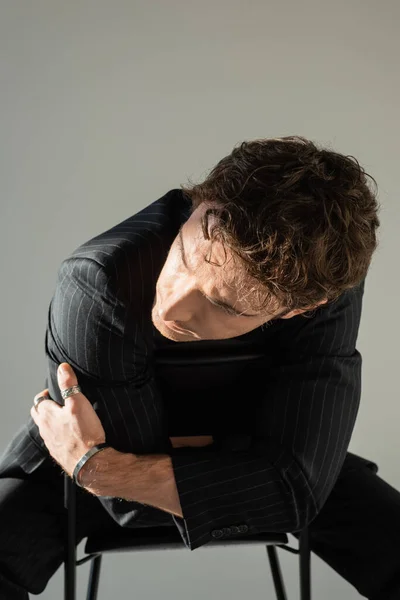 Brunette man in black striped blazer and metal bracelet posing sitting on chair with crossed arms isolated on grey — Stock Photo