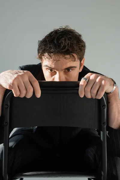 Brunette man in black clothes hiding face behind chair back and looking at camera isolated on grey — Foto stock