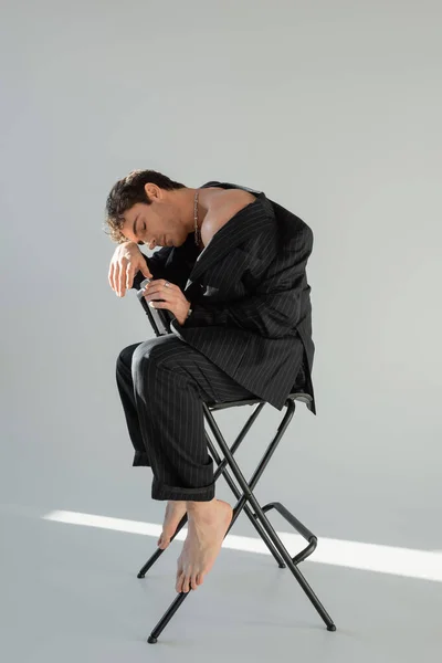 Full length of sexy barefoot man with closed eyes sitting on chair in black and striped suit on grey background - foto de stock