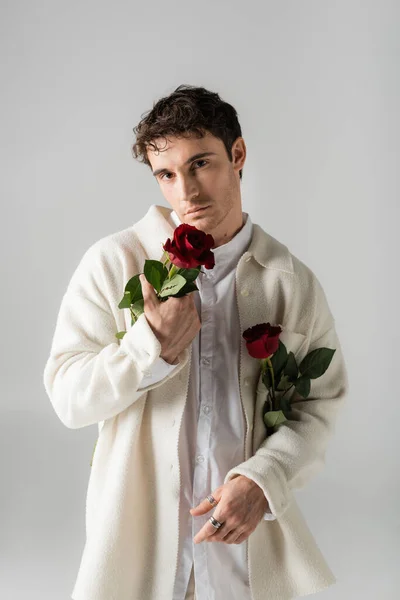 Brunette man in white soft jacket and shirt holding red roses and looking at camera isolated on grey - foto de stock